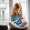 Red haired charming schoolgirl in blue dress browsing smartphone while sitting on rustic wooden table with legs crossed beside books against big window at home